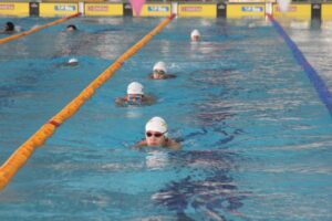 Young swimmers in a pool swimming breaststroke during a swim meet, wearing goggles and swim caps, surrounded by lane dividers.