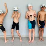 Four young swimmers wearing swim caps and competitive swimwear stand in a playful pose against a plain wall.