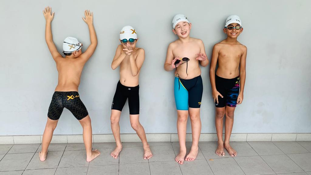 Four young swimmers wearing swim caps and competitive swimwear stand in a playful pose against a plain wall.