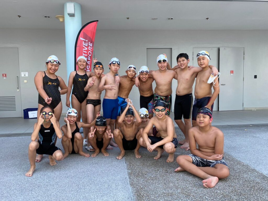 A group of young swimmers in swimwear and caps posing together outdoors near a poolside, smiling and making cheerful gestures.