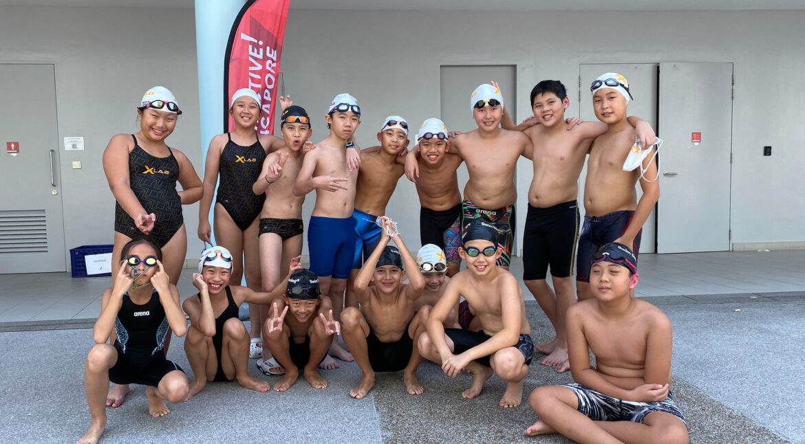 A group of young swimmers in swimwear and caps posing together outdoors near a poolside, smiling and making cheerful gestures.