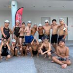 A group of young swimmers in swimwear and caps posing together outdoors near a poolside, smiling and making cheerful gestures.